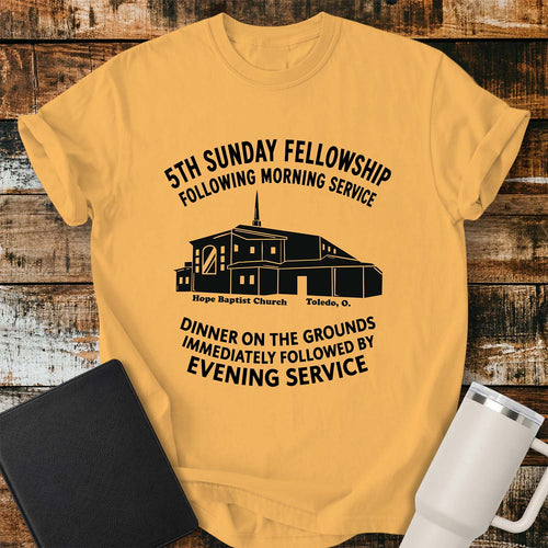 citrus yellow T-shirt featuring a black graphic of Hope Baptist Church with event text reading “5TH SUNDAY FELLOWSHIP FOLLOWING MORNING SERVICE” and “DINNER ON THE GROUNDS IMMEDIATELY FOLLOWED BY EVENING SERVICE.” The church illustration is labeled “Hope Baptist Church, Toledo, O.” The shirt is displayed on a rustic wood plank background alongside a black Bible and a white insulated tumbler.