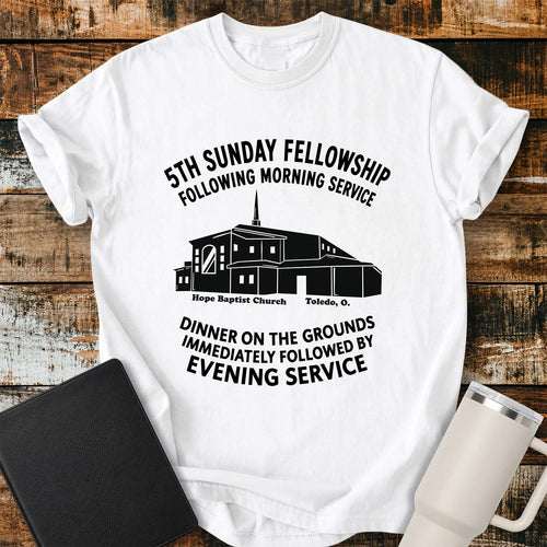 White T-shirt featuring a black graphic of Hope Baptist Church with event text reading “5TH SUNDAY FELLOWSHIP FOLLOWING MORNING SERVICE” and “DINNER ON THE GROUNDS IMMEDIATELY FOLLOWED BY EVENING SERVICE.” The church illustration is labeled “Hope Baptist Church, Toledo, O.” The shirt is displayed on a rustic wood plank background alongside a black Bible and a white insulated tumbler.
