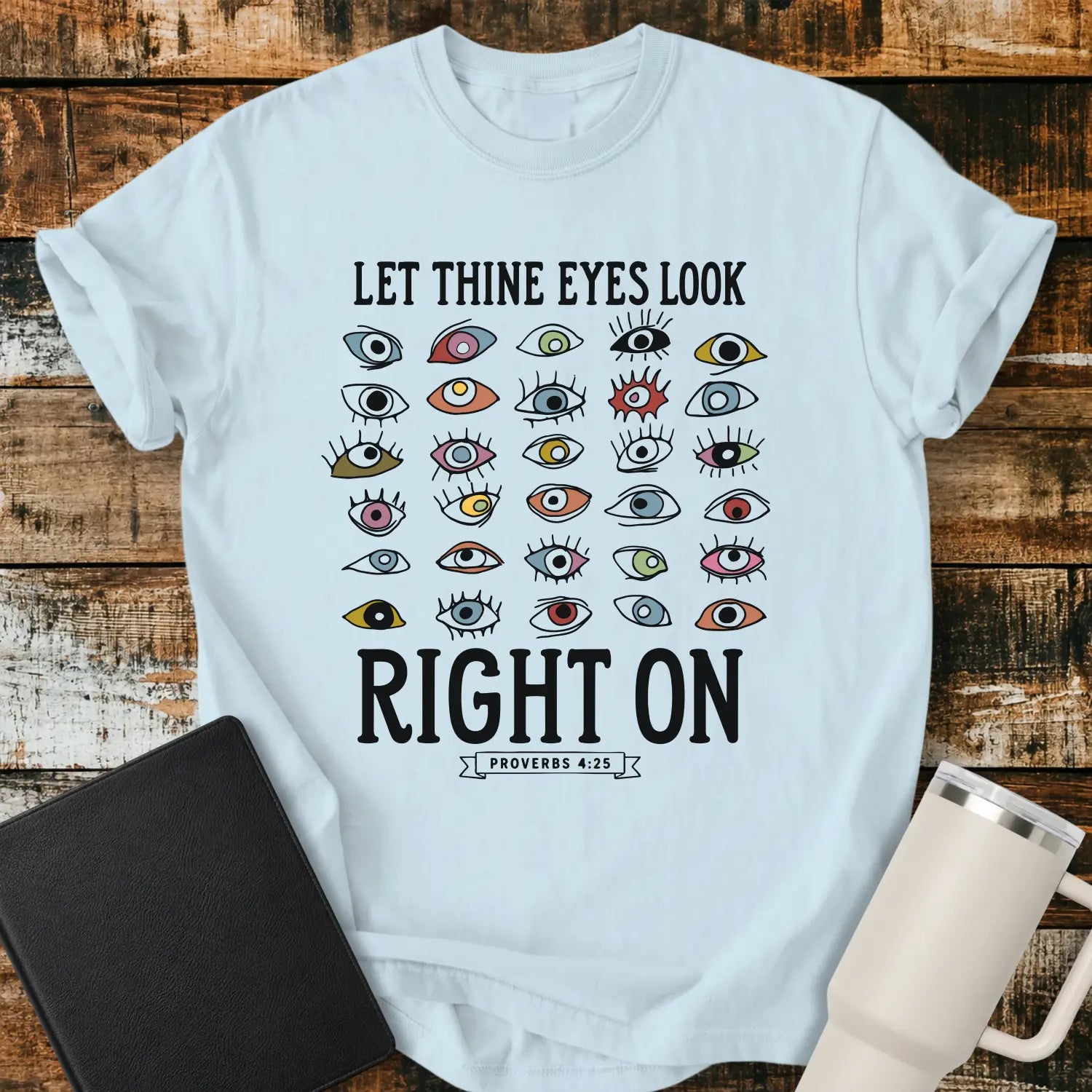 Chambray blue graphic T-shirt mockup featuring the phrase “Let Thine Eyes Look Right On” with a Proverbs 4:25 reference, surrounded by a collection of colorful illustrated eyes, styled with a black Bible and white tumbler on a rustic wood background.