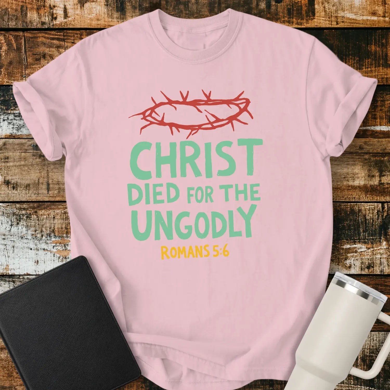 blossom pink T-shirt mockup featuring the Christian message “CHRIST DIED FOR THE UNGODLY” with Romans 5:6 beneath in contrasting lettering. A red crown of thorns graphic appears above the text, emphasizing the gospel theme. The shirt is displayed on a rustic wood plank background beside a black Bible and a white insulated tumbler, creating a faith-based apparel presentation.