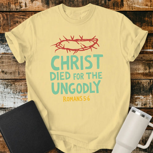 butter yellow T-shirt mockup featuring the Christian message “CHRIST DIED FOR THE UNGODLY” with Romans 5:6 beneath in contrasting lettering. A red crown of thorns graphic appears above the text, emphasizing the gospel theme. The shirt is displayed on a rustic wood plank background beside a black Bible and a white insulated tumbler, creating a faith-based apparel presentation.