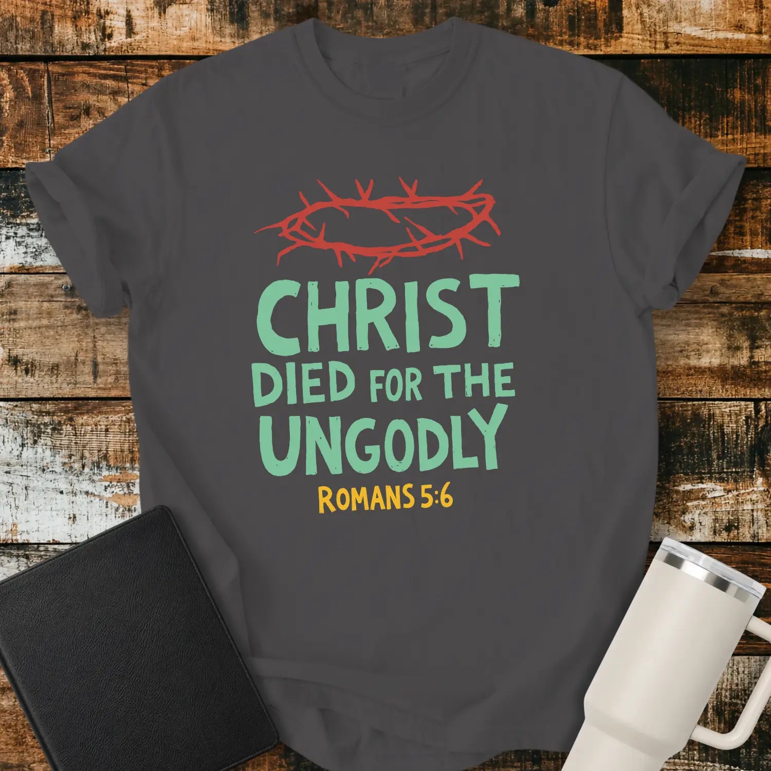 graphite T-shirt mockup featuring the Christian message “CHRIST DIED FOR THE UNGODLY” with Romans 5:6 beneath in contrasting lettering. A red crown of thorns graphic appears above the text, emphasizing the gospel theme. The shirt is displayed on a rustic wood plank background beside a black Bible and a white insulated tumbler, creating a faith-based apparel presentation.