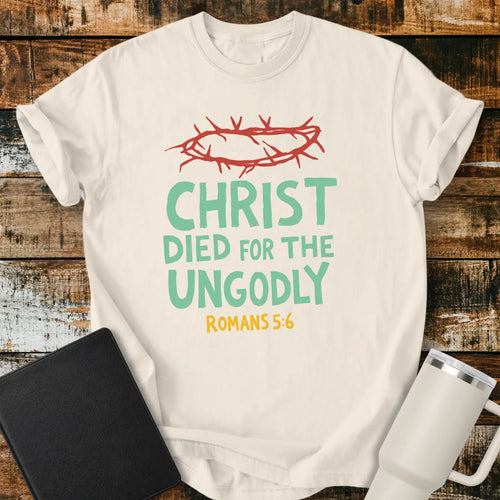 ivory T-shirt mockup featuring the Christian message “CHRIST DIED FOR THE UNGODLY” with Romans 5:6 beneath in contrasting lettering. A red crown of thorns graphic appears above the text, emphasizing the gospel theme. The shirt is displayed on a rustic wood plank background beside a black Bible and a white insulated tumbler, creating a faith-based apparel presentation.