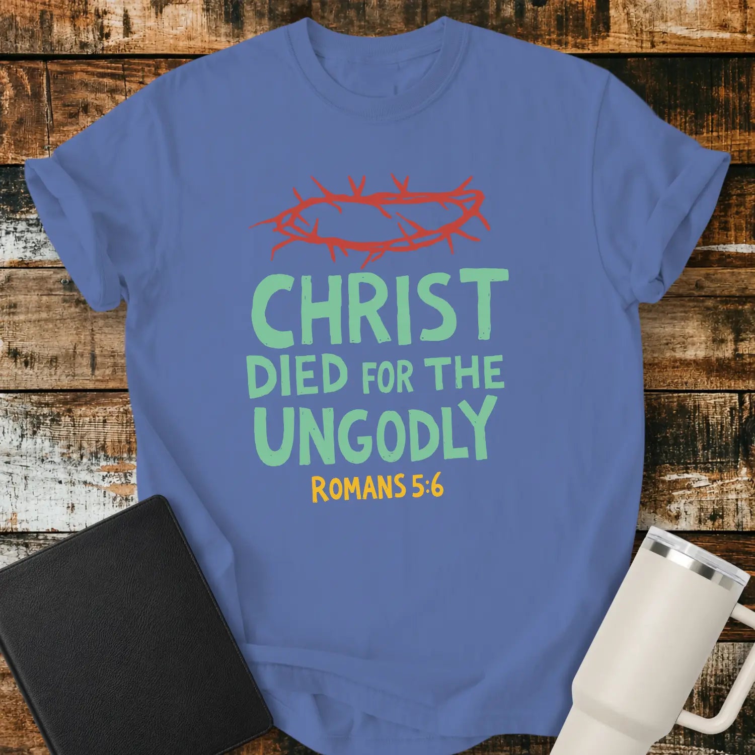 mystic blue T-shirt mockup featuring the Christian message “CHRIST DIED FOR THE UNGODLY” with Romans 5:6 beneath in contrasting lettering. A red crown of thorns graphic appears above the text, emphasizing the gospel theme. The shirt is displayed on a rustic wood plank background beside a black Bible and a white insulated tumbler, creating a faith-based apparel presentation.