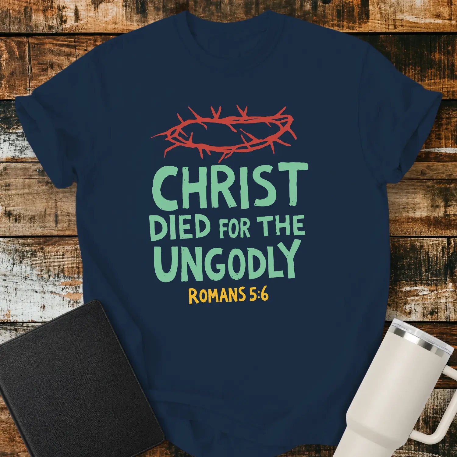 True Navy T-shirt mockup featuring the Christian message “CHRIST DIED FOR THE UNGODLY” with Romans 5:6 beneath in contrasting lettering. A red crown of thorns graphic appears above the text, emphasizing the gospel theme. The shirt is displayed on a rustic wood plank background beside a black Bible and a white insulated tumbler, creating a faith-based apparel presentation.
