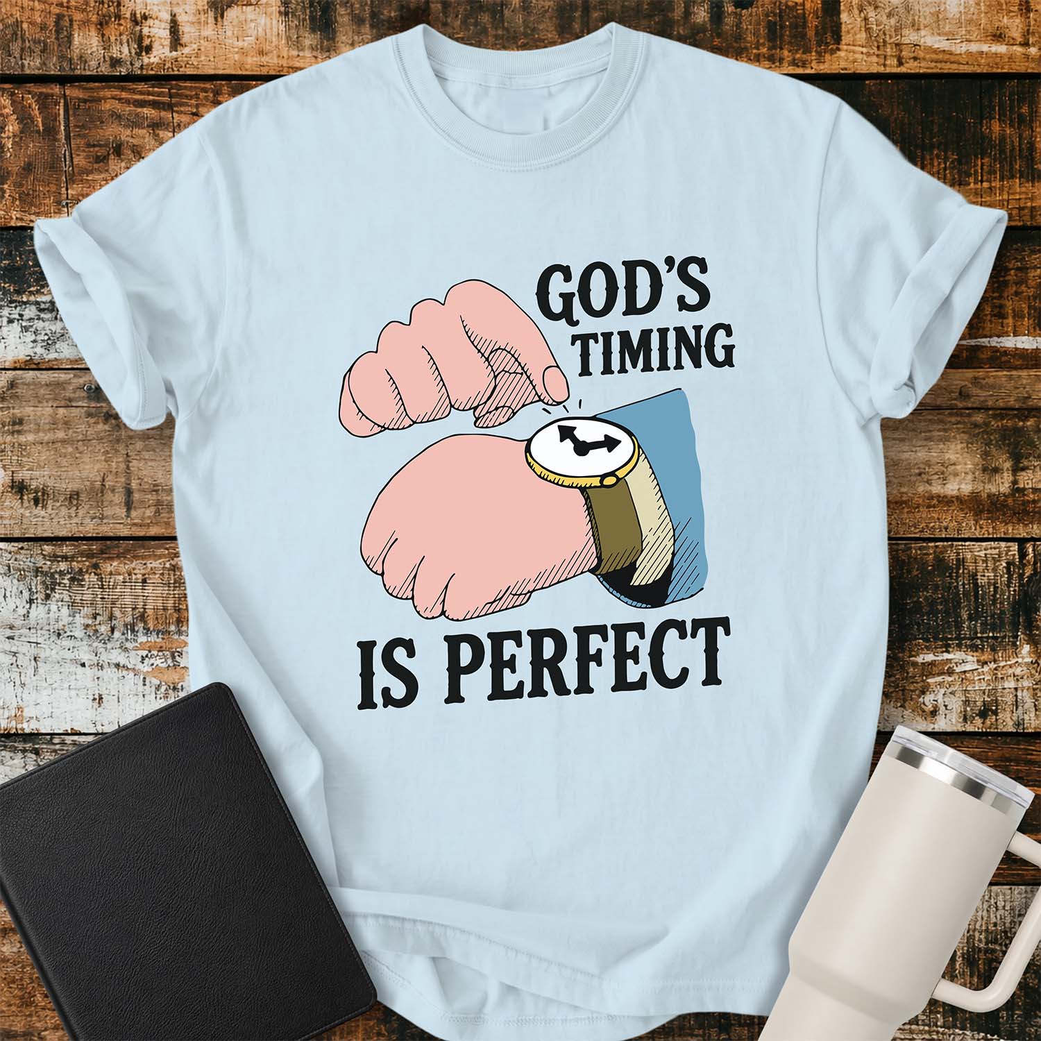 Chambray blue T-shirt featuring a colorful illustration of a hand tapping a wristwatch beside the phrase “GOD’S TIMING IS PERFECT” in bold black lettering. The watch has a gold bezel, brown band, and white face with black hands, reinforcing the message about trusting God's timing. The shirt is displayed on a rustic wood plank background with a black Bible on the left and a white insulated tumbler on the right.