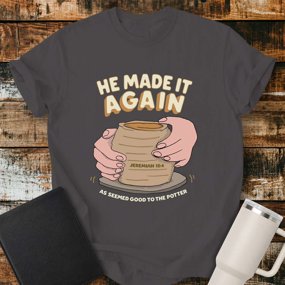 graphite Christian t-shirt featuring hands shaping a clay vessel on a potter’s wheel, with the phrase “He Made It Again,” Jeremiah 18:4, and “As Seemed Good to the Potter.” A retro-inspired biblical design illustrating God as the Potter.