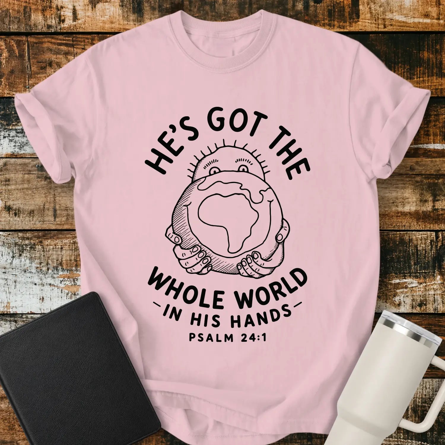 blossom pink mockup featuring the phrase “HE’S GOT THE WHOLE WORLD IN HIS HANDS” with Psalm 24:1 beneath. The design shows a cheerful figure holding a globe, illustrated in a simple vintage line-art style. The shirt is displayed on a rustic wood plank background beside a black Bible and a white insulated tumbler, creating a faith-based apparel presentation.