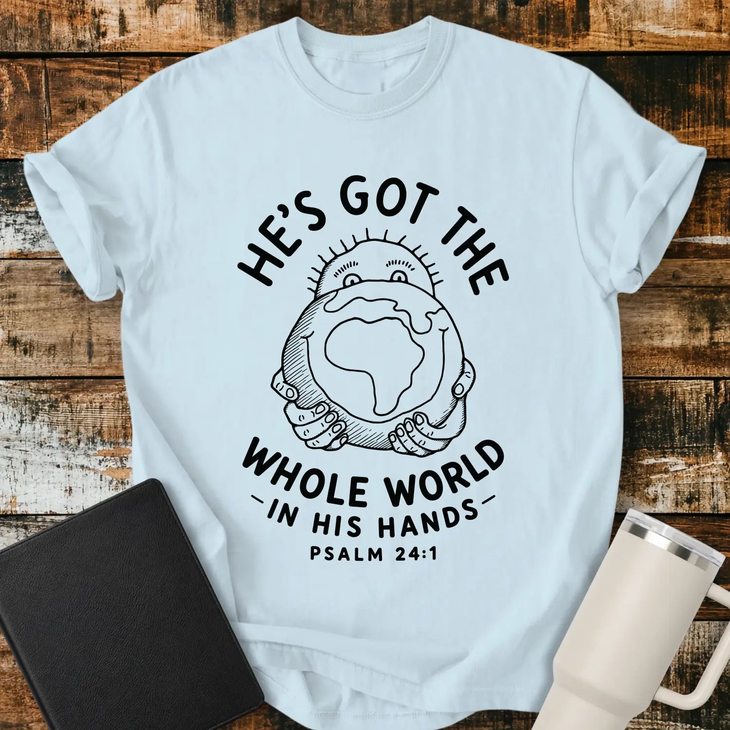 chambray blue mockup featuring the phrase “HE’S GOT THE WHOLE WORLD IN HIS HANDS” with Psalm 24:1 beneath. The design shows a cheerful figure holding a globe, illustrated in a simple vintage line-art style. The shirt is displayed on a rustic wood plank background beside a black Bible and a white insulated tumbler, creating a faith-based apparel presentation.