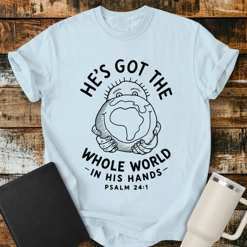 chambray blue mockup featuring the phrase “HE’S GOT THE WHOLE WORLD IN HIS HANDS” with Psalm 24:1 beneath. The design shows a cheerful figure holding a globe, illustrated in a simple vintage line-art style. The shirt is displayed on a rustic wood plank background beside a black Bible and a white insulated tumbler, creating a faith-based apparel presentation.