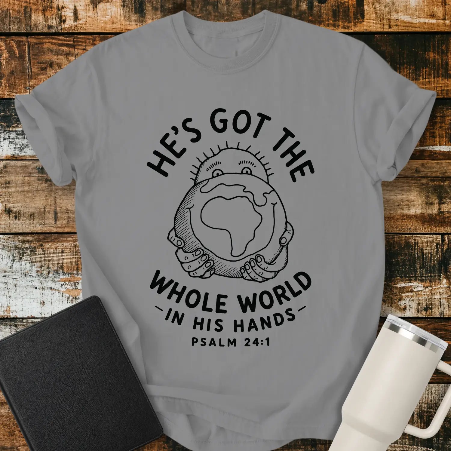 grey mockup featuring the phrase “HE’S GOT THE WHOLE WORLD IN HIS HANDS” with Psalm 24:1 beneath. The design shows a cheerful figure holding a globe, illustrated in a simple vintage line-art style. The shirt is displayed on a rustic wood plank background beside a black Bible and a white insulated tumbler, creating a faith-based apparel presentation.