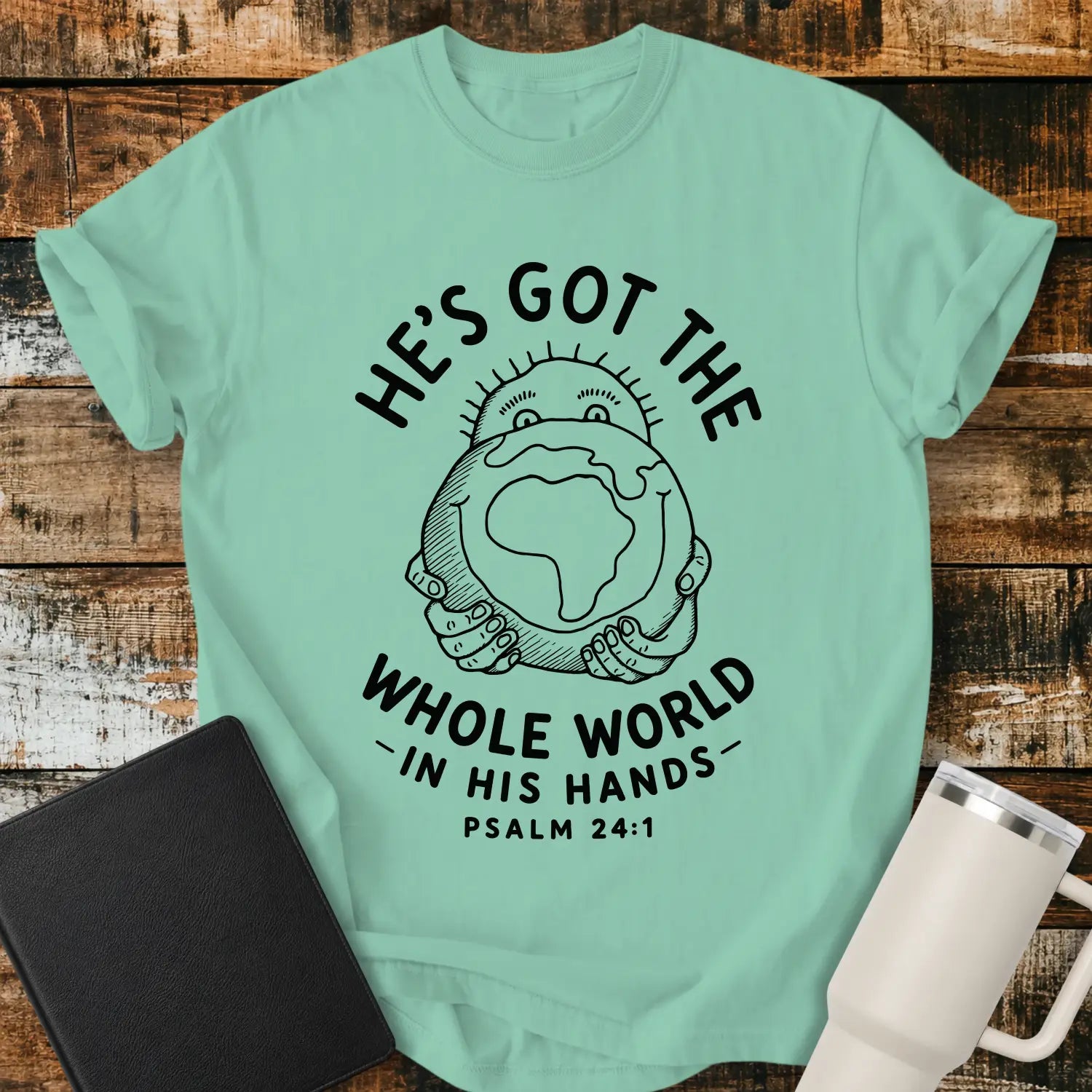 Island Green mockup featuring the phrase “HE’S GOT THE WHOLE WORLD IN HIS HANDS” with Psalm 24:1 beneath. The design shows a cheerful figure holding a globe, illustrated in a simple vintage line-art style. The shirt is displayed on a rustic wood plank background beside a black Bible and a white insulated tumbler, creating a faith-based apparel presentation.
