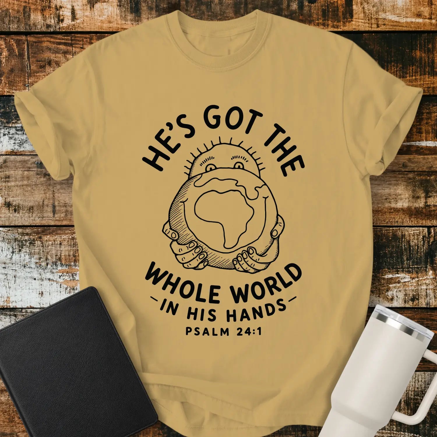 mustard yellow mockup featuring the phrase “HE’S GOT THE WHOLE WORLD IN HIS HANDS” with Psalm 24:1 beneath. The design shows a cheerful figure holding a globe, illustrated in a simple vintage line-art style. The shirt is displayed on a rustic wood plank background beside a black Bible and a white insulated tumbler, creating a faith-based apparel presentation.
