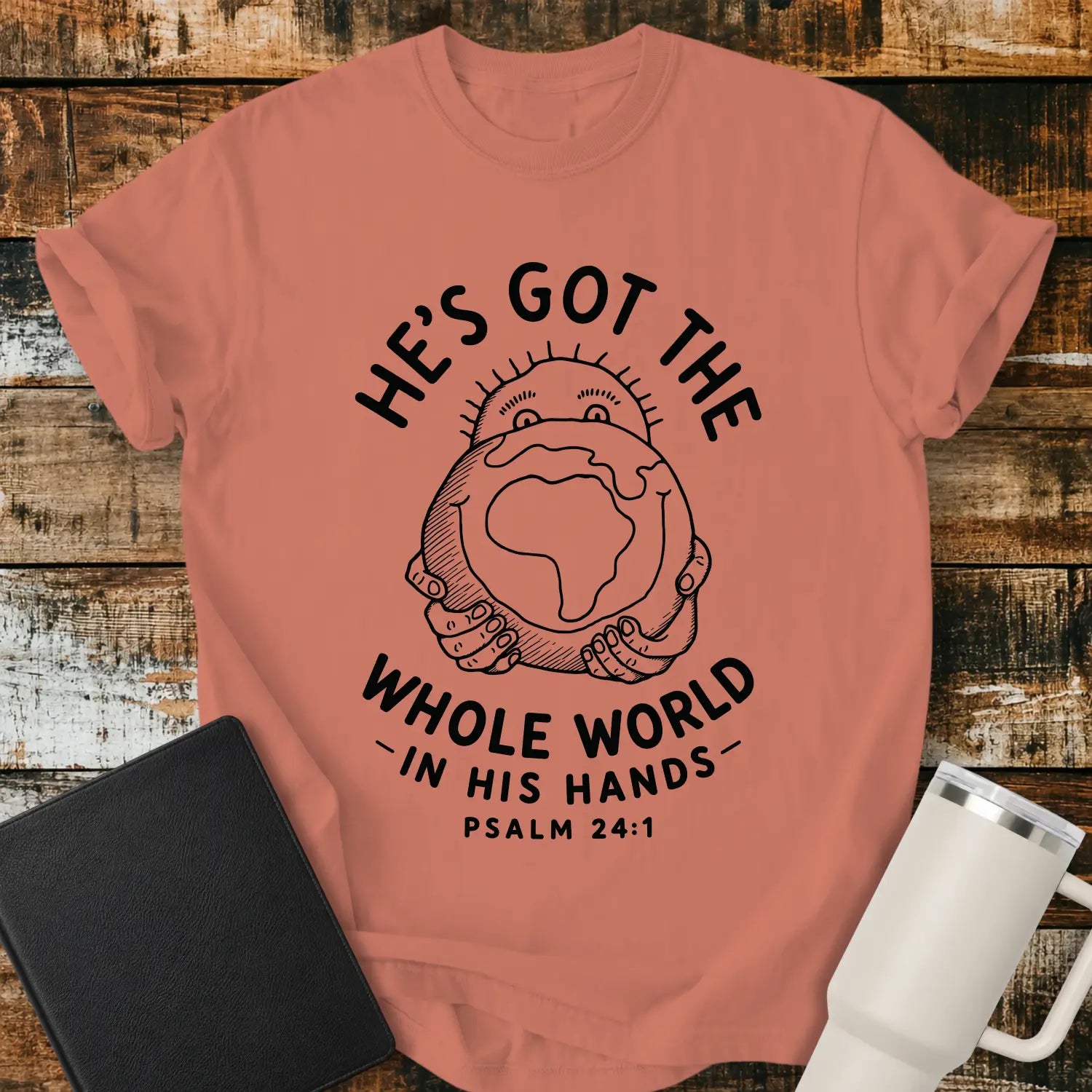 terracotta mockup featuring the phrase “HE’S GOT THE WHOLE WORLD IN HIS HANDS” with Psalm 24:1 beneath. The design shows a cheerful figure holding a globe, illustrated in a simple vintage line-art style. The shirt is displayed on a rustic wood plank background beside a black Bible and a white insulated tumbler, creating a faith-based apparel presentation.