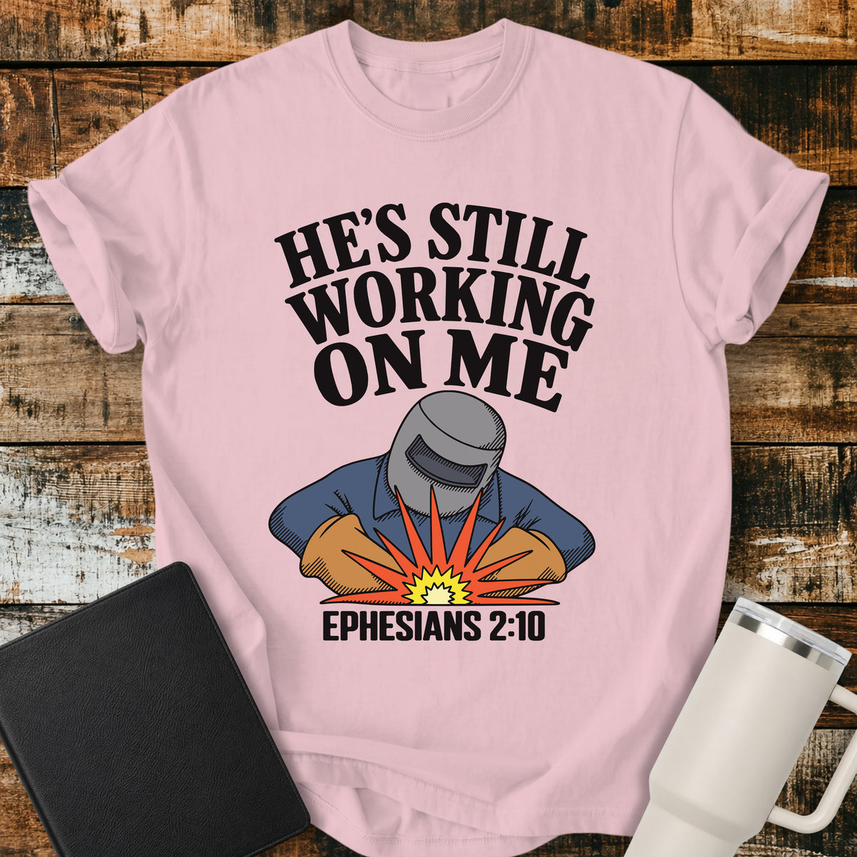 blossom pink Christian t-shirt featuring a vintage welder illustration with bold retro lettering reading “He’s Still Working on Me” and Ephesians 2:10.