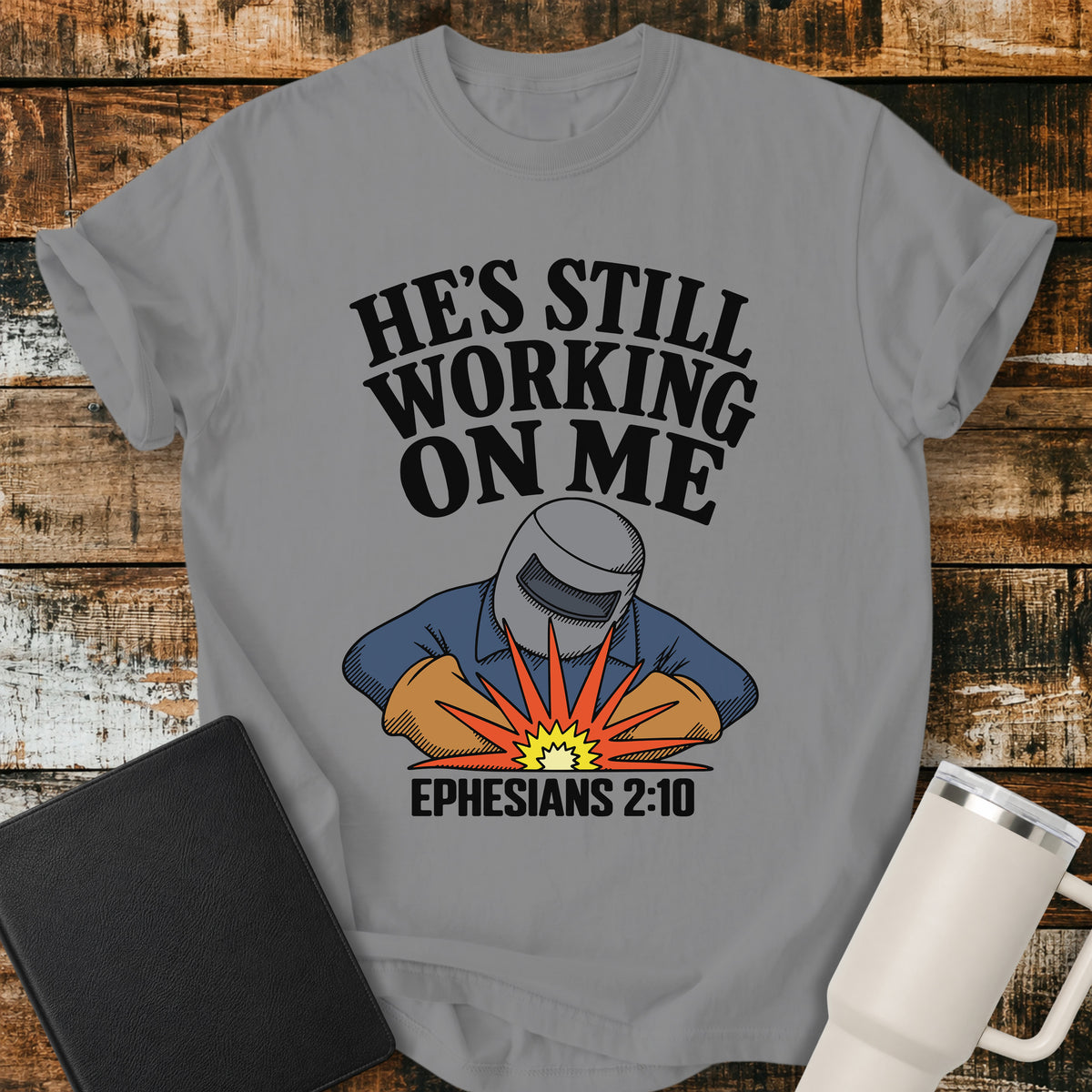 grey Christian t-shirt featuring a vintage welder illustration with bold retro lettering reading “He’s Still Working on Me” and Ephesians 2:10.