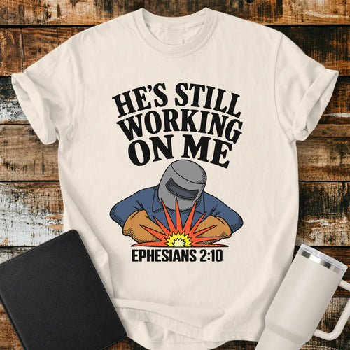 Ivory Christian t-shirt featuring a vintage welder illustration with bold retro lettering reading “He’s Still Working on Me” and Ephesians 2:10.