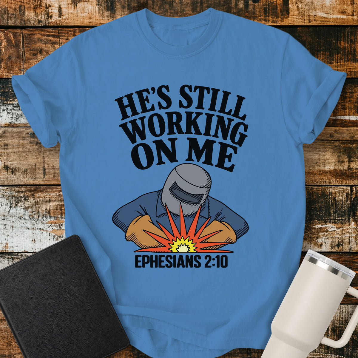 royal caribe blue Christian t-shirt featuring a vintage welder illustration with bold retro lettering reading “He’s Still Working on Me” and Ephesians 2:10.