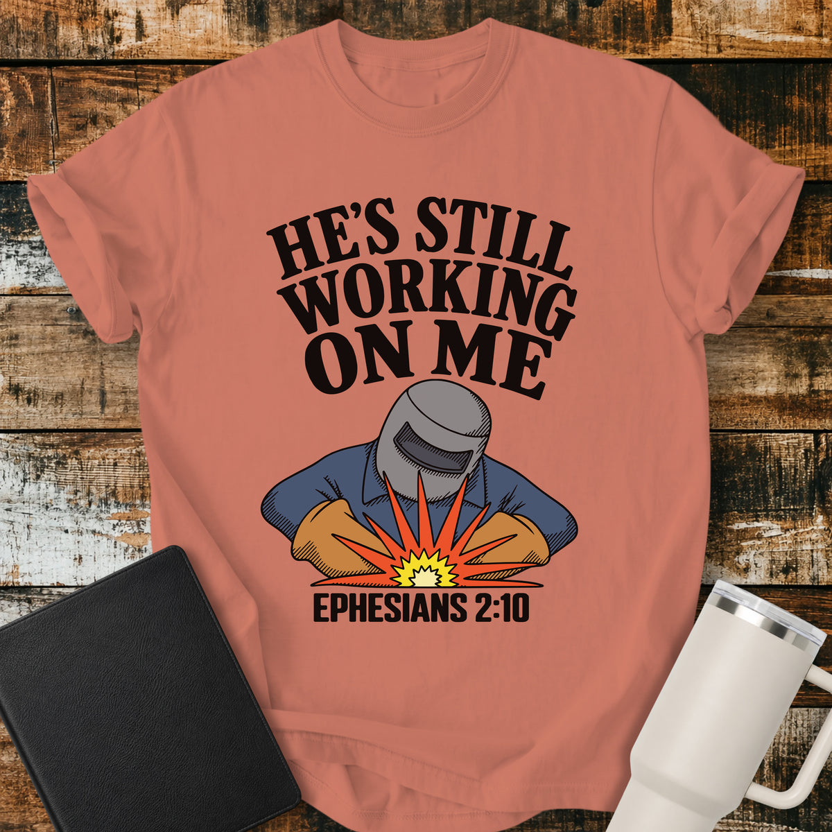 terracotta Christian t-shirt featuring a vintage welder illustration with bold retro lettering reading “He’s Still Working on Me” and Ephesians 2:10.