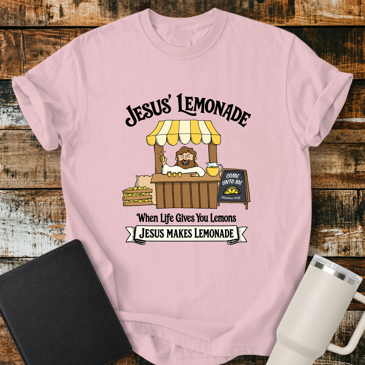 blossom pink flat-lay t-shirt mockup featuring a cheerful illustration of Jesus serving lemonade from a vintage lemonade stand. The design includes the phrases "JESUS' LEMONADE," "When Life Gives You Lemons," and "JESUS MAKES LEMONADE." A chalkboard sign beside the stand reads, "COME UNTO ME" with the Bible reference "Matthew 11:28." The shirt is displayed on a rustic wood plank background alongside a black leather Bible and a white insulated travel mug.