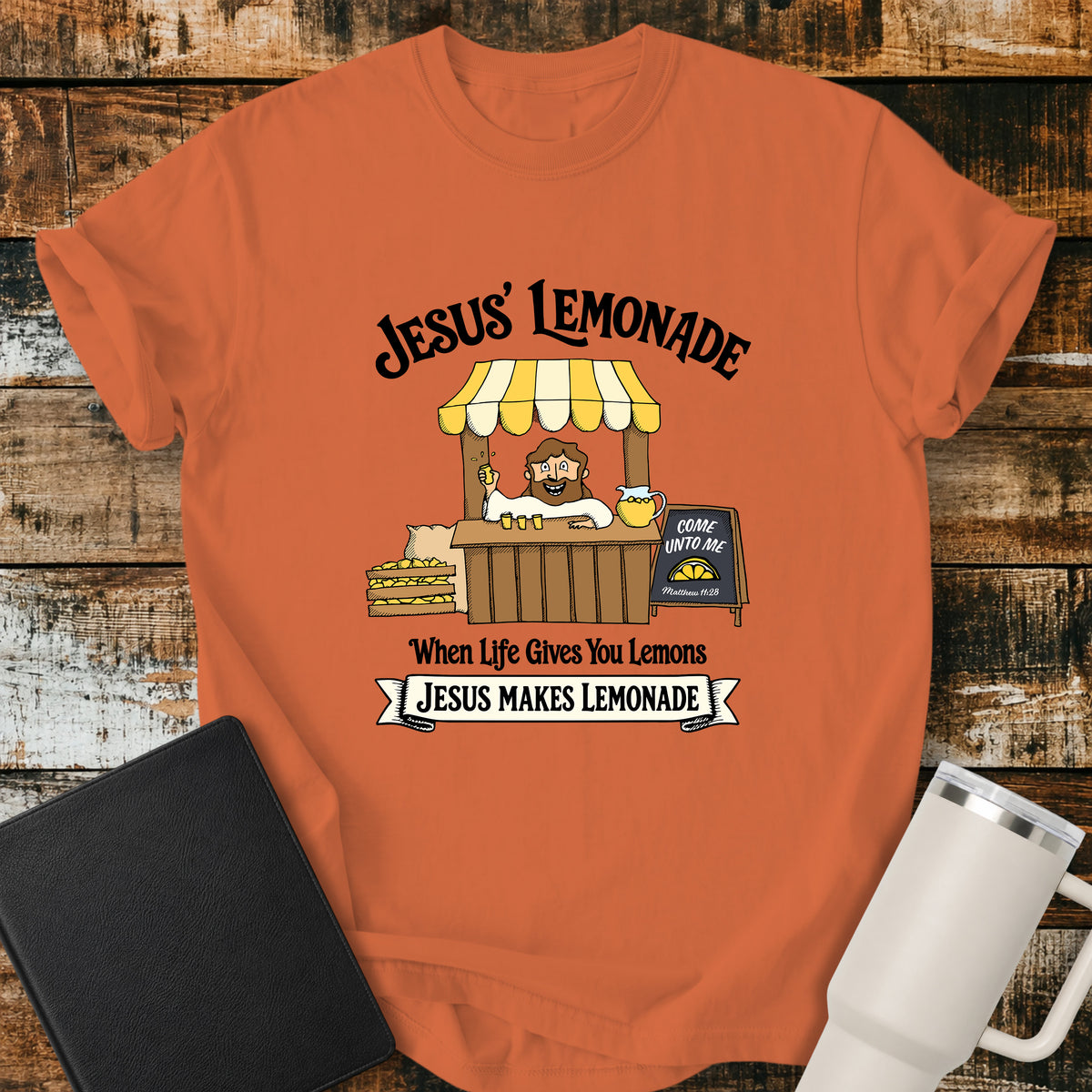 burnt orange flat-lay t-shirt mockup featuring a cheerful illustration of Jesus serving lemonade from a vintage lemonade stand. The design includes the phrases "JESUS' LEMONADE," "When Life Gives You Lemons," and "JESUS MAKES LEMONADE." A chalkboard sign beside the stand reads, "COME UNTO ME" with the Bible reference "Matthew 11:28." The shirt is displayed on a rustic wood plank background alongside a black leather Bible and a white insulated travel mug.