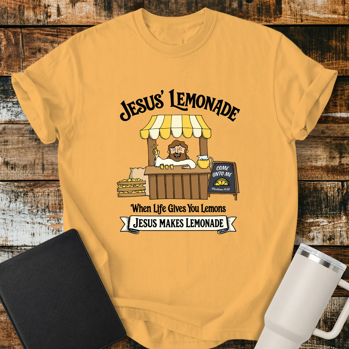 Citrus yellow flat-lay t-shirt mockup featuring a cheerful illustration of Jesus serving lemonade from a vintage lemonade stand. The design includes the phrases "JESUS' LEMONADE," "When Life Gives You Lemons," and "JESUS MAKES LEMONADE." A chalkboard sign beside the stand reads, "COME UNTO ME" with the Bible reference "Matthew 11:28." The shirt is displayed on a rustic wood plank background alongside a black leather Bible and a white insulated travel mug.