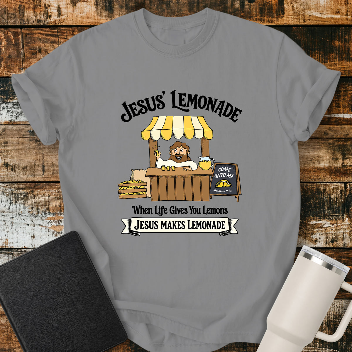 grey flat-lay t-shirt mockup featuring a cheerful illustration of Jesus serving lemonade from a vintage lemonade stand. The design includes the phrases "JESUS' LEMONADE," "When Life Gives You Lemons," and "JESUS MAKES LEMONADE." A chalkboard sign beside the stand reads, "COME UNTO ME" with the Bible reference "Matthew 11:28." The shirt is displayed on a rustic wood plank background alongside a black leather Bible and a white insulated travel mug.