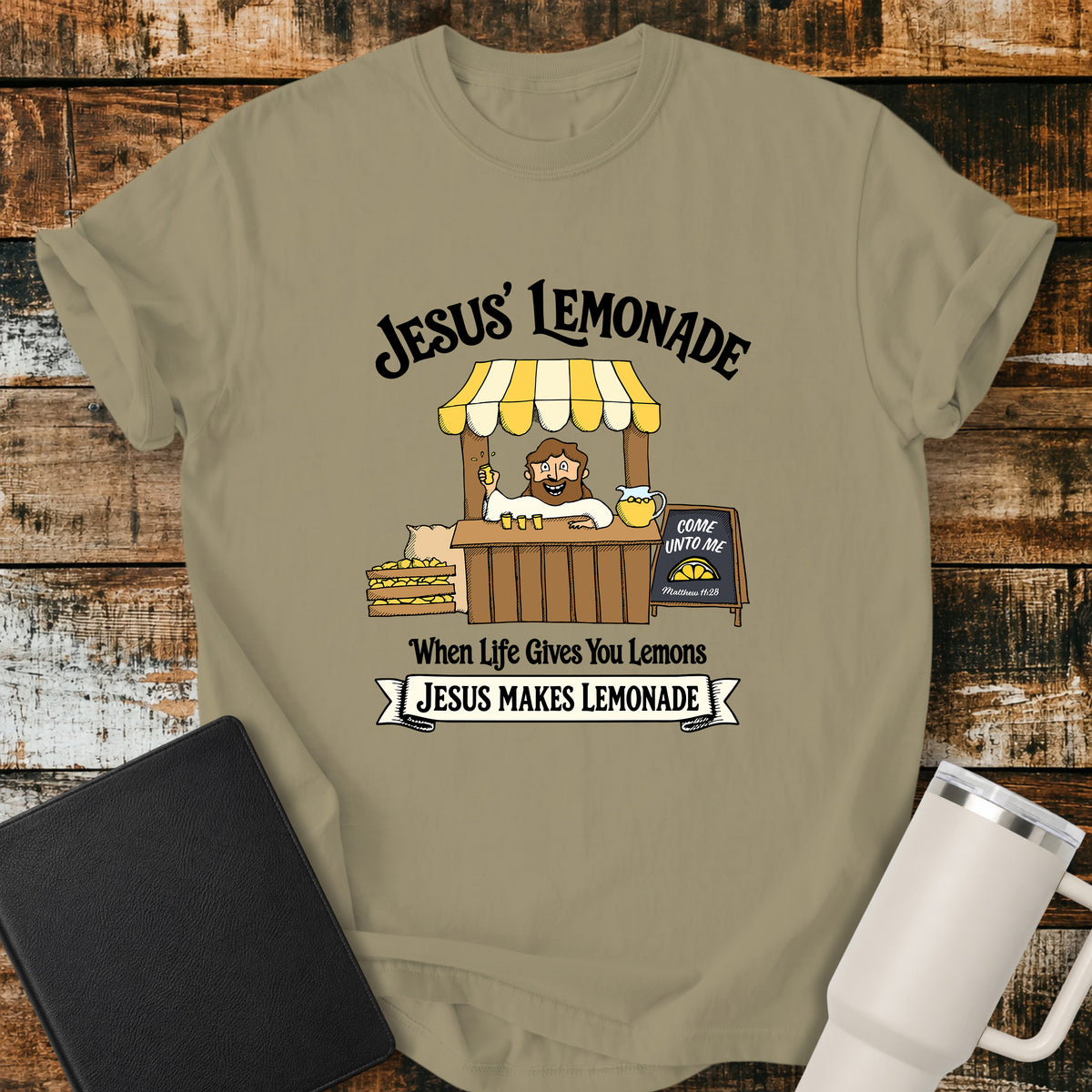 khaki flat-lay t-shirt mockup featuring a cheerful illustration of Jesus serving lemonade from a vintage lemonade stand. The design includes the phrases "JESUS' LEMONADE," "When Life Gives You Lemons," and "JESUS MAKES LEMONADE." A chalkboard sign beside the stand reads, "COME UNTO ME" with the Bible reference "Matthew 11:28." The shirt is displayed on a rustic wood plank background alongside a black leather Bible and a white insulated travel mug.