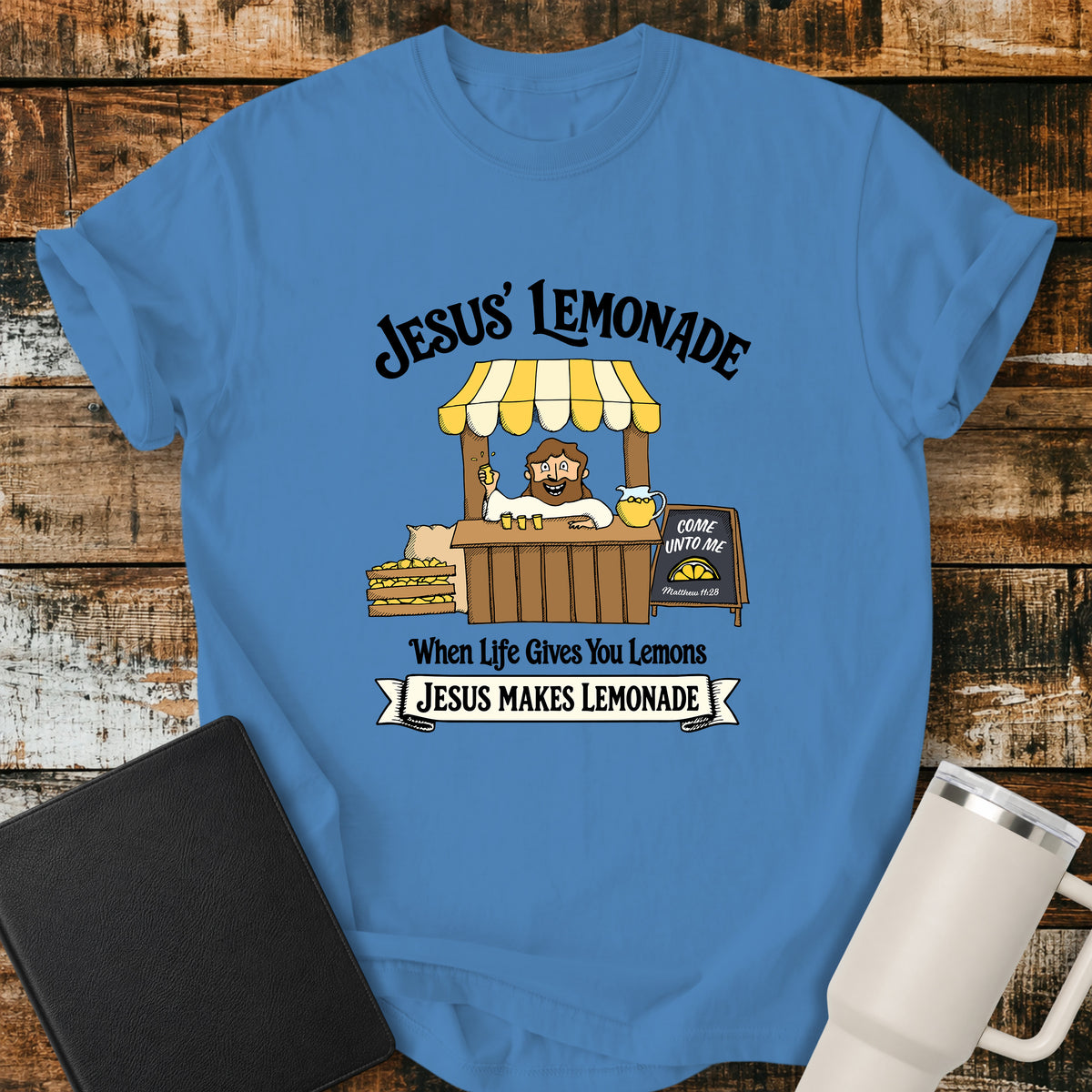 royal caribe blue flat-lay t-shirt mockup featuring a cheerful illustration of Jesus serving lemonade from a vintage lemonade stand. The design includes the phrases "JESUS' LEMONADE," "When Life Gives You Lemons," and "JESUS MAKES LEMONADE." A chalkboard sign beside the stand reads, "COME UNTO ME" with the Bible reference "Matthew 11:28." The shirt is displayed on a rustic wood plank background alongside a black leather Bible and a white insulated travel mug.