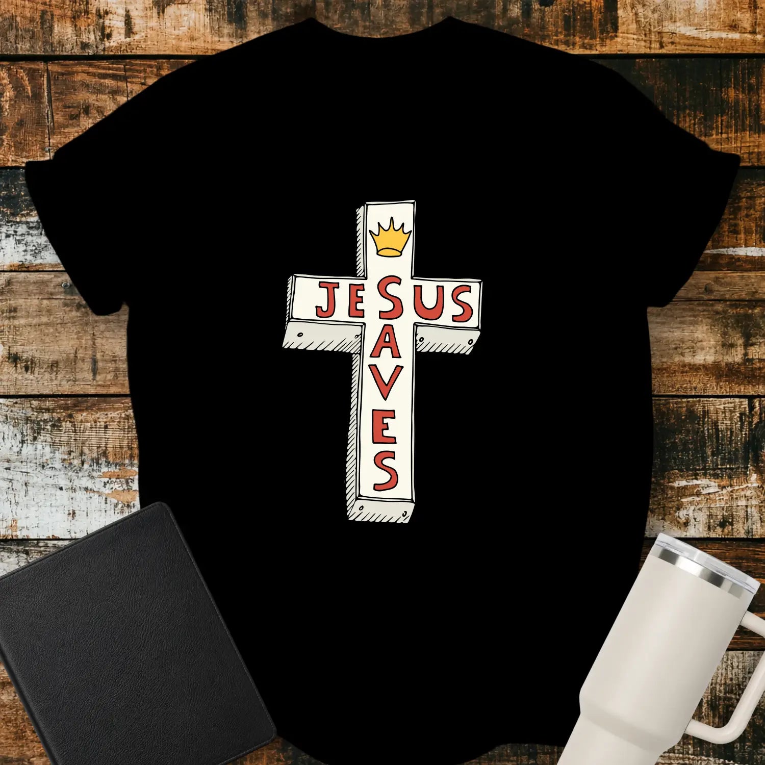 black T-shirt mockup featuring a white cross graphic with the words “JESUS SAVES” integrated into the cross design in red lettering and a small gold crown at the top. The shirt is displayed on a rustic wood background alongside a black Bible and a white insulated tumbler.
