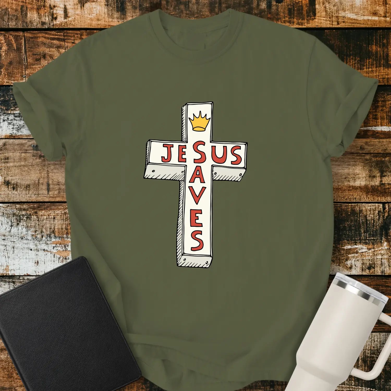 hemp green T-shirt mockup featuring a white cross graphic with the words “JESUS SAVES” integrated into the cross design in red lettering and a small gold crown at the top. The shirt is displayed on a rustic wood background alongside a black Bible and a white insulated tumbler.
