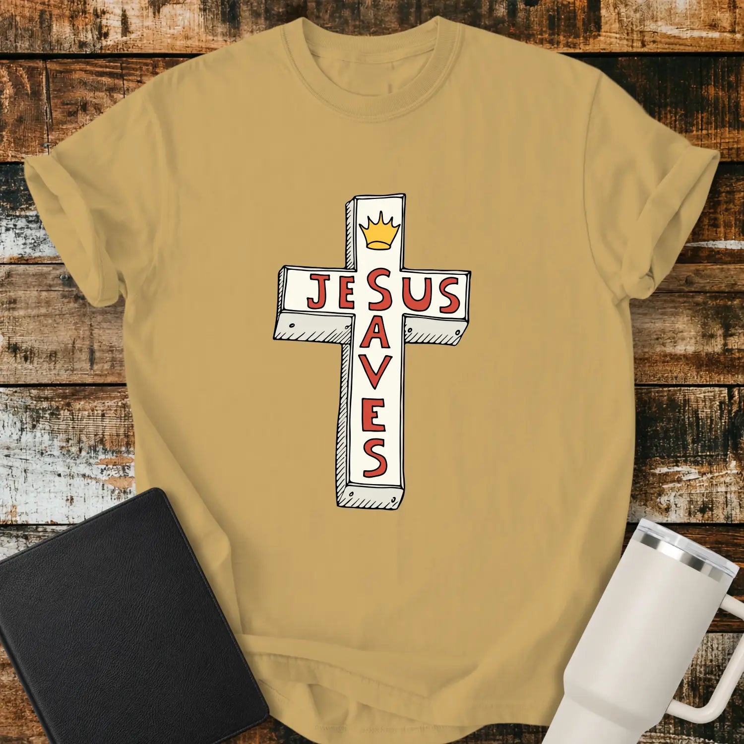 Mustard yellow T-shirt mockup featuring a white cross graphic with the words “JESUS SAVES” integrated into the cross design in red lettering and a small gold crown at the top. The shirt is displayed on a rustic wood background alongside a black Bible and a white insulated tumbler.
