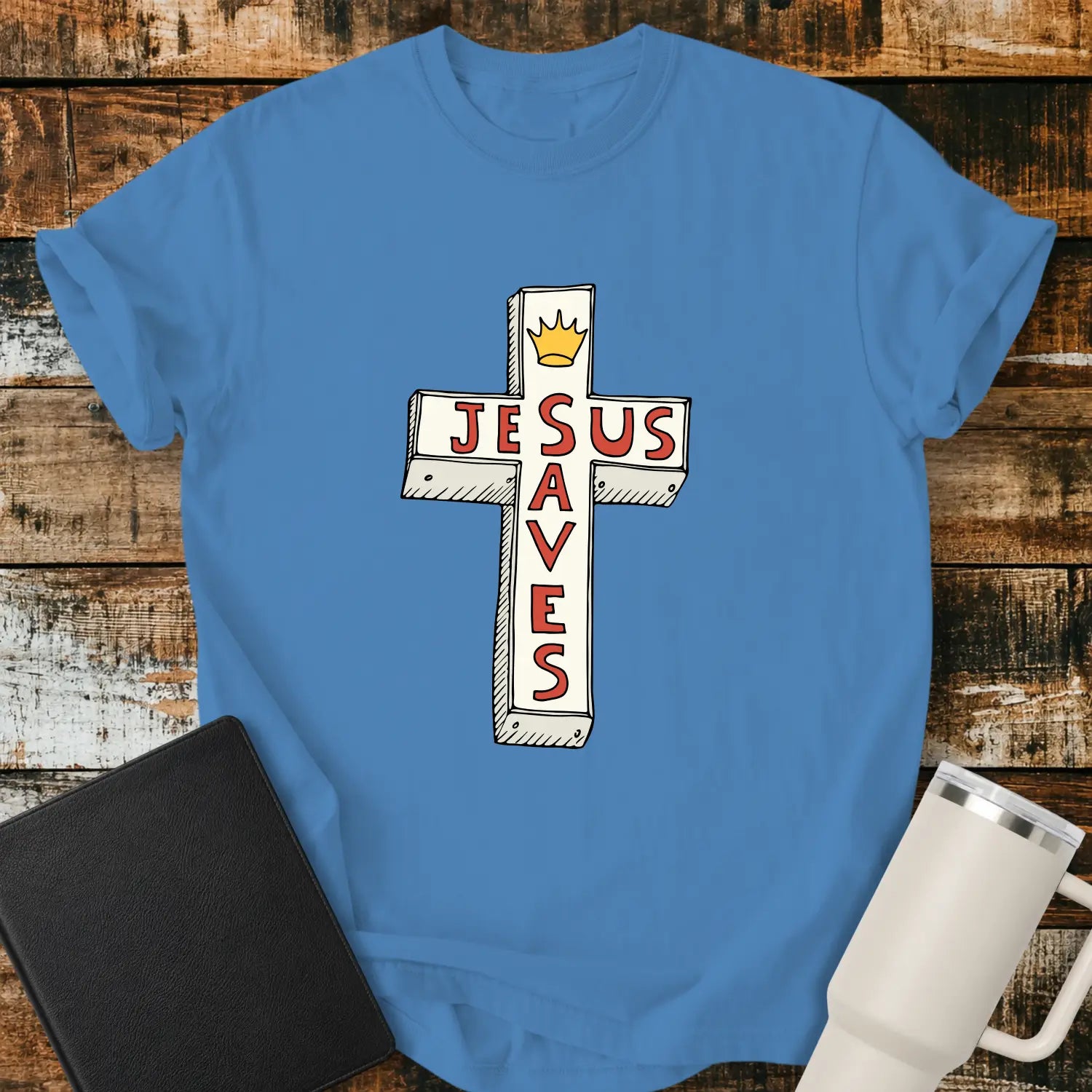 royal caribe blue T-shirt mockup featuring a white cross graphic with the words “JESUS SAVES” integrated into the cross design in red lettering and a small gold crown at the top. The shirt is displayed on a rustic wood background alongside a black Bible and a white insulated tumbler.
