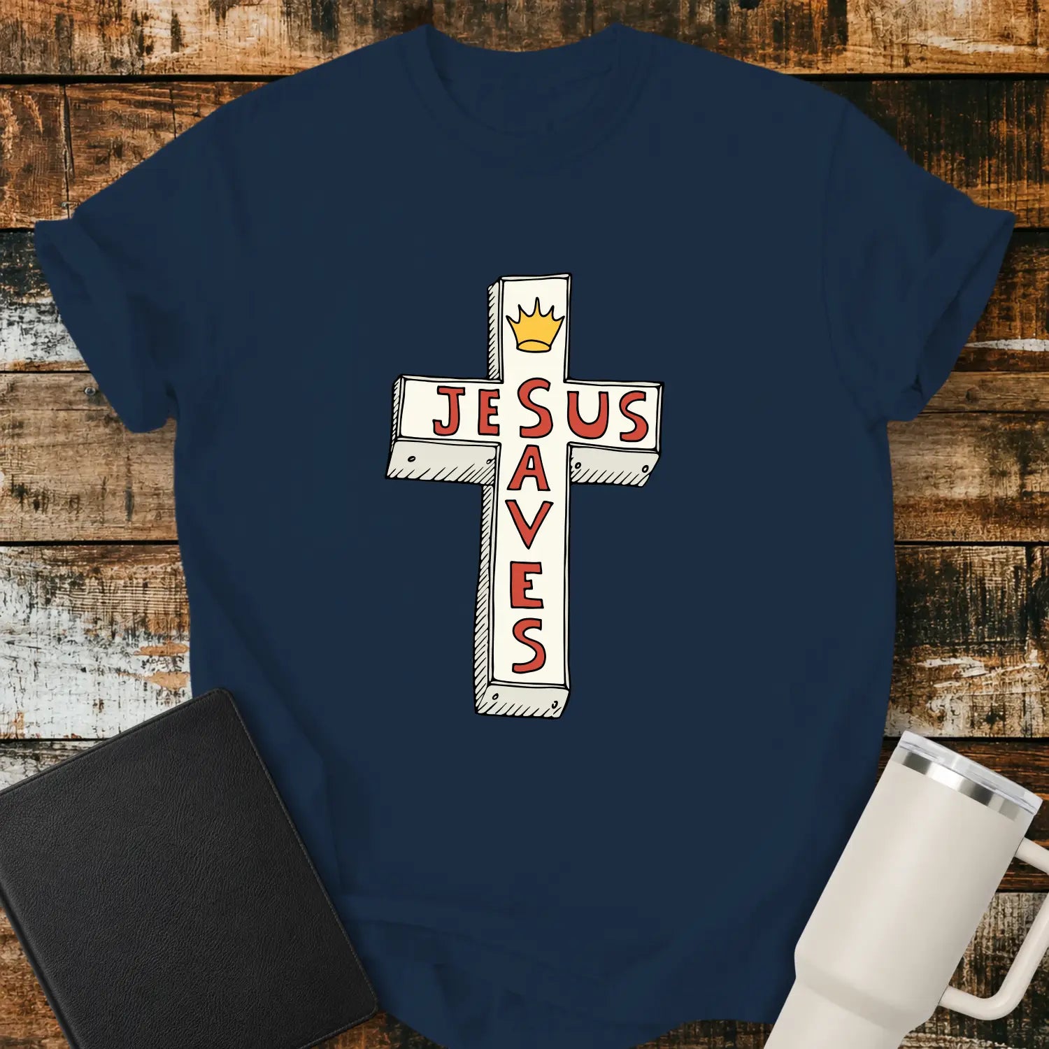 true navy T-shirt mockup featuring a white cross graphic with the words “JESUS SAVES” integrated into the cross design in red lettering and a small gold crown at the top. The shirt is displayed on a rustic wood background alongside a black Bible and a white insulated tumbler.
