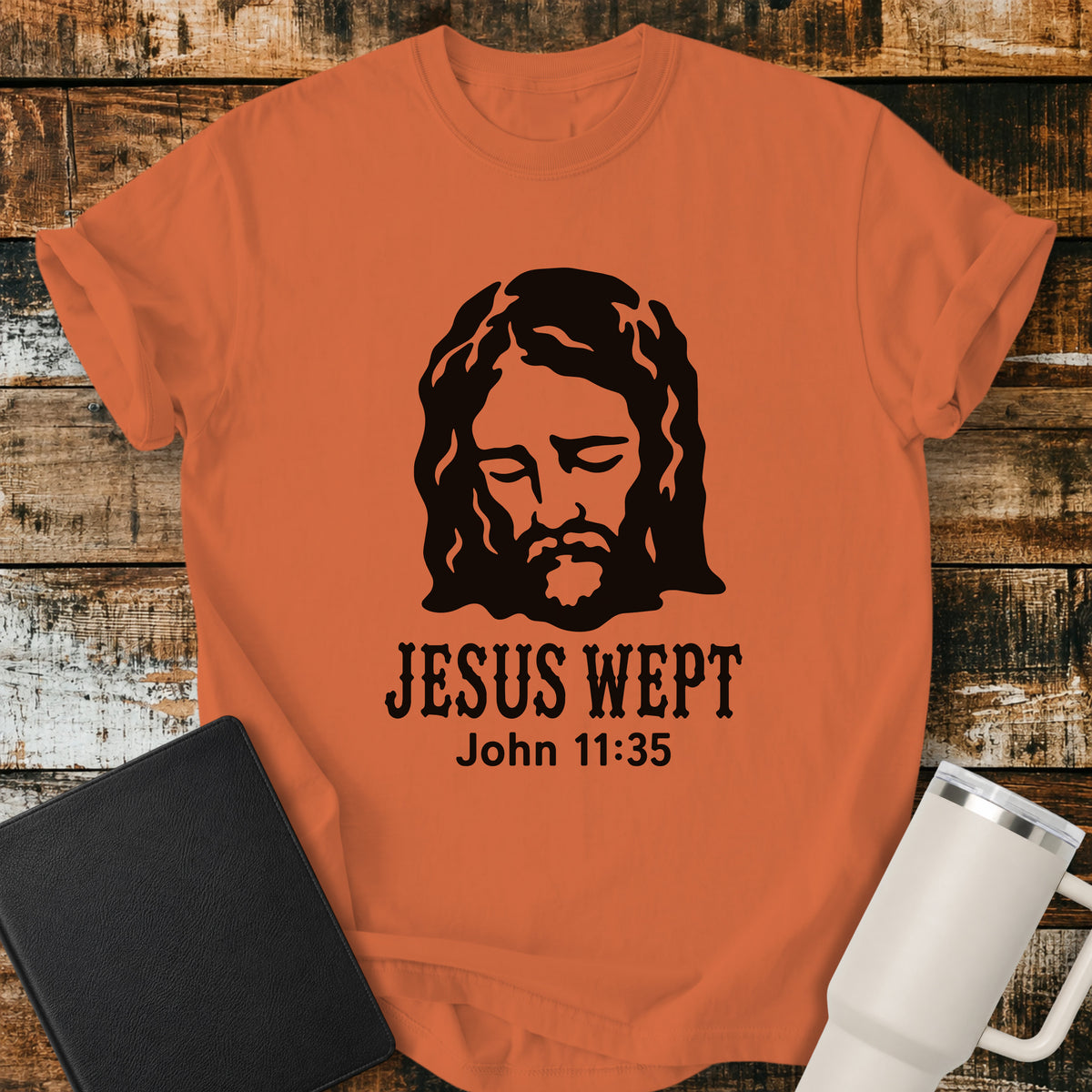 burnt orange flat-lay t-shirt mockup featuring a minimalist black illustration of Jesus with His head bowed and eyes closed above the words, "JESUS WEPT," with the Bible reference "John 11:35" below. The simple faith-inspired design is displayed on a rustic wood plank background alongside a black leather Bible and a white insulated travel mug.