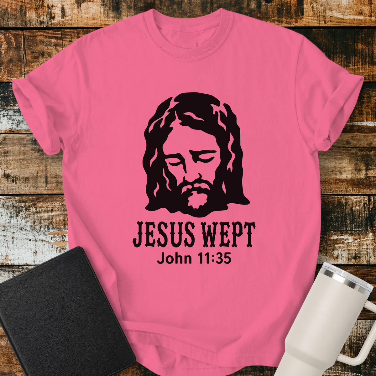 crunchberry pink flat-lay t-shirt mockup featuring a minimalist black illustration of Jesus with His head bowed and eyes closed above the words, "JESUS WEPT," with the Bible reference "John 11:35" below. The simple faith-inspired design is displayed on a rustic wood plank background alongside a black leather Bible and a white insulated travel mug.