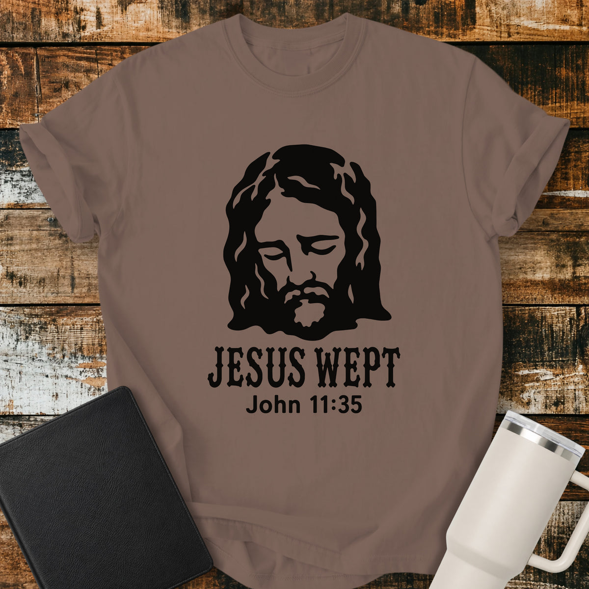 expresso brown flat-lay t-shirt mockup featuring a minimalist black illustration of Jesus with His head bowed and eyes closed above the words, "JESUS WEPT," with the Bible reference "John 11:35" below. The simple faith-inspired design is displayed on a rustic wood plank background alongside a black leather Bible and a white insulated travel mug.