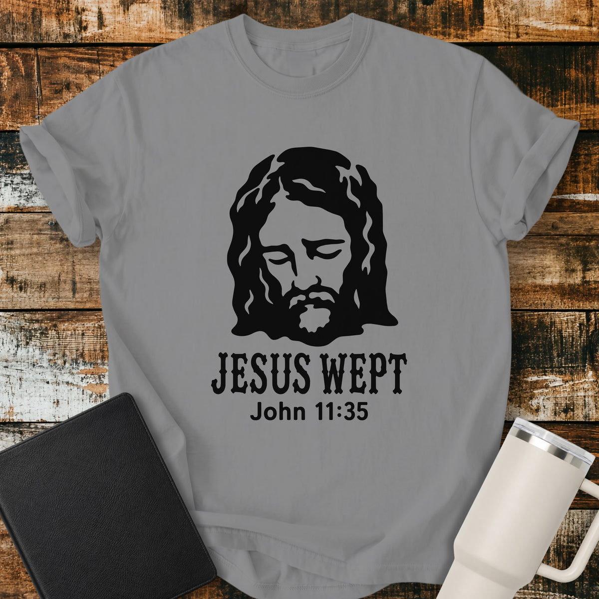 grey flat-lay t-shirt mockup featuring a minimalist black illustration of Jesus with His head bowed and eyes closed above the words, "JESUS WEPT," with the Bible reference "John 11:35" below. The simple faith-inspired design is displayed on a rustic wood plank background alongside a black leather Bible and a white insulated travel mug.