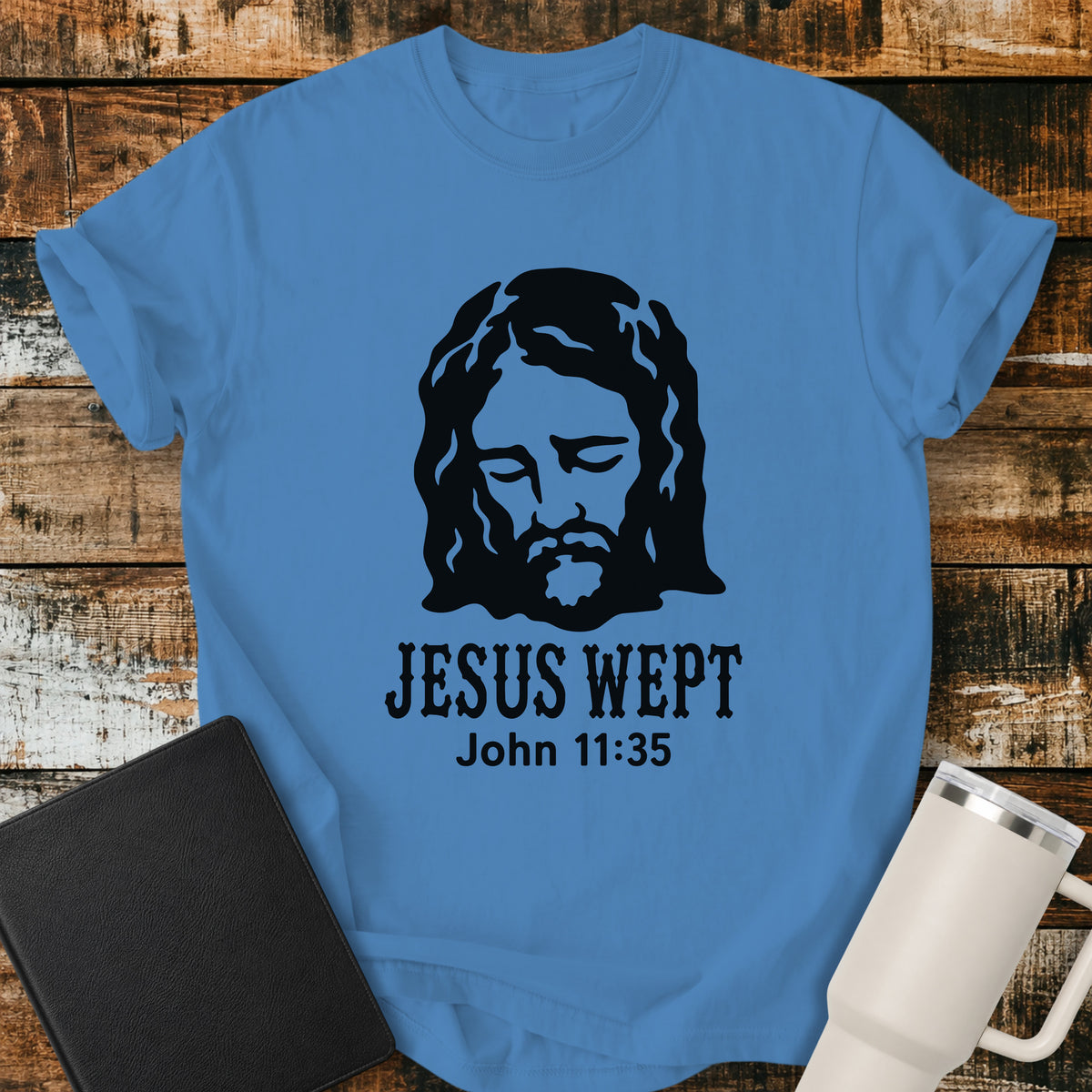 royal caribe blue flat-lay t-shirt mockup featuring a minimalist black illustration of Jesus with His head bowed and eyes closed above the words, "JESUS WEPT," with the Bible reference "John 11:35" below. The simple faith-inspired design is displayed on a rustic wood plank background alongside a black leather Bible and a white insulated travel mug.