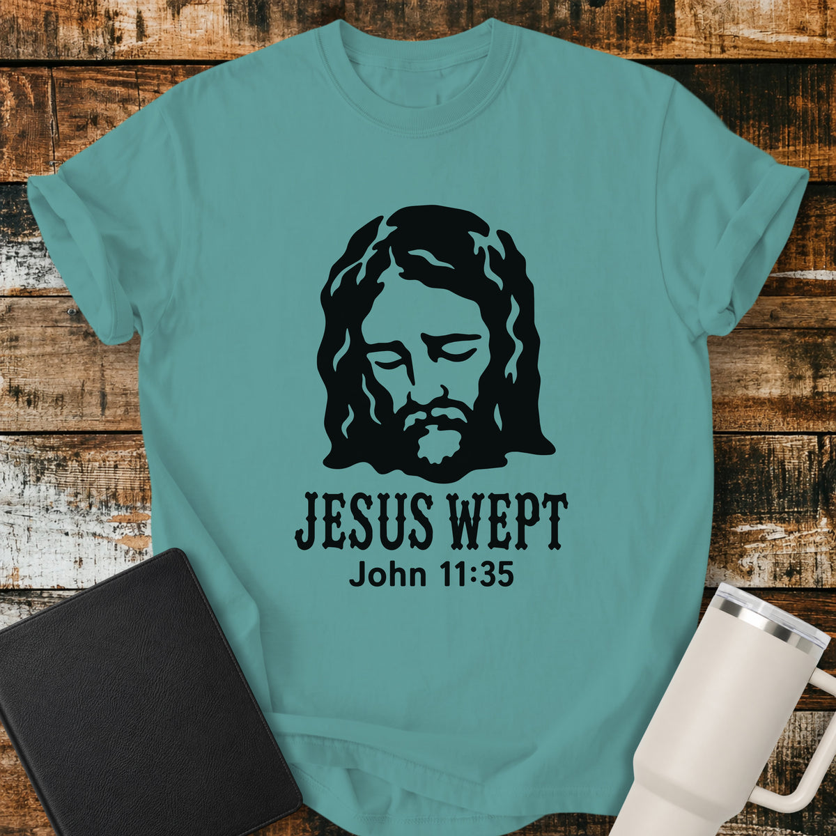 seafoam green flat-lay t-shirt mockup featuring a minimalist black illustration of Jesus with His head bowed and eyes closed above the words, "JESUS WEPT," with the Bible reference "John 11:35" below. The simple faith-inspired design is displayed on a rustic wood plank background alongside a black leather Bible and a white insulated travel mug.