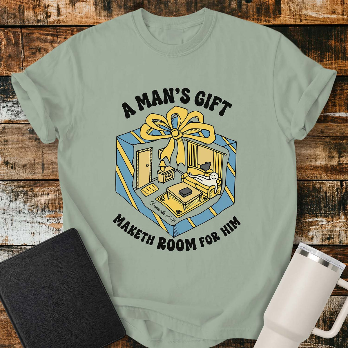 bay green flat-lay t-shirt mockup featuring a colorful retro illustration of a gift box opened into a cozy living room with a sofa, lamp, coffee table, door, and window beneath a large yellow bow. Curved black lettering reads, "A MAN'S GIFT" above and "MAKETH ROOM FOR HIM" below, inspired by Proverbs 18:16. The shirt is displayed on a rustic wood plank background with a black leather Bible and a white insulated travel mug.