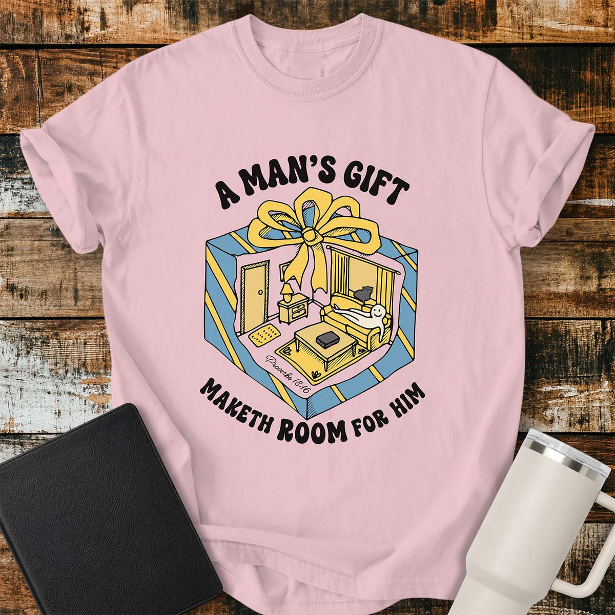 blossom pink flat-lay t-shirt mockup featuring a colorful retro illustration of a gift box opened into a cozy living room with a sofa, lamp, coffee table, door, and window beneath a large yellow bow. Curved black lettering reads, "A MAN'S GIFT" above and "MAKETH ROOM FOR HIM" below, inspired by Proverbs 18:16. The shirt is displayed on a rustic wood plank background with a black leather Bible and a white insulated travel mug.