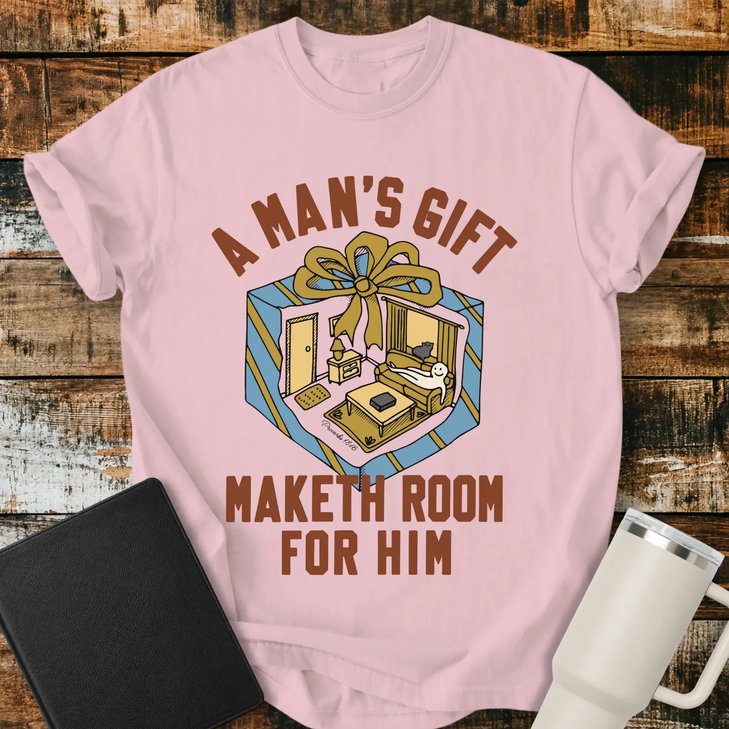 blossom pink graphic T-shirt mockup featuring the phrase “A Man’s Gift Maketh Room for Him” in vintage-style lettering with an illustration of a gift box opening into a furnished room scene, styled with a black notebook and white tumbler on a rustic wood background.