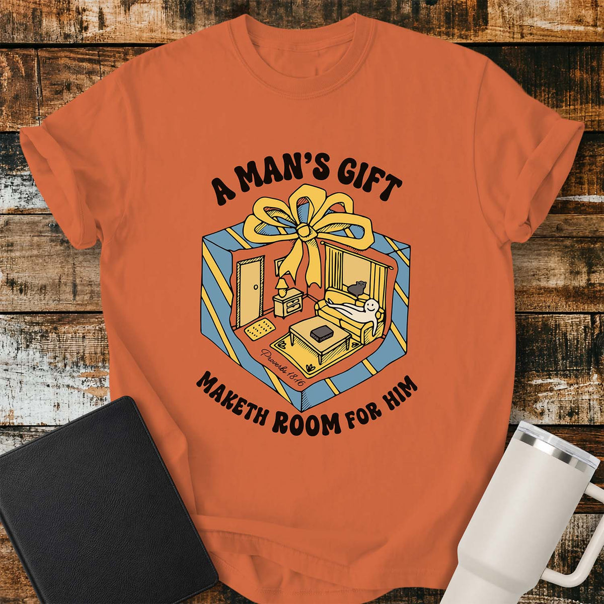 Burnt orange flat-lay t-shirt mockup featuring a colorful retro illustration of a gift box opened into a cozy living room with a sofa, lamp, coffee table, door, and window beneath a large yellow bow. Curved black lettering reads, "A MAN'S GIFT" above and "MAKETH ROOM FOR HIM" below, inspired by Proverbs 18:16. The shirt is displayed on a rustic wood plank background with a black leather Bible and a white insulated travel mug.