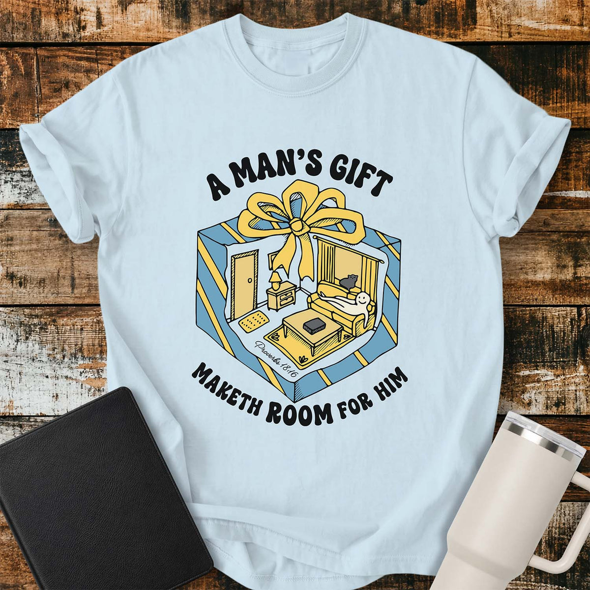 chambray blue flat-lay t-shirt mockup featuring a colorful retro illustration of a gift box opened into a cozy living room with a sofa, lamp, coffee table, door, and window beneath a large yellow bow. Curved black lettering reads, "A MAN'S GIFT" above and "MAKETH ROOM FOR HIM" below, inspired by Proverbs 18:16. The shirt is displayed on a rustic wood plank background with a black leather Bible and a white insulated travel mug.