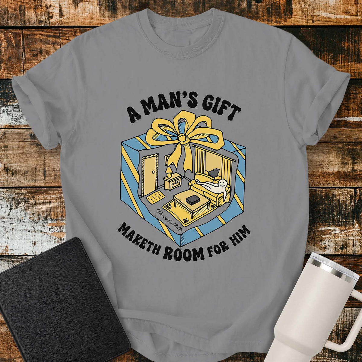 grey flat-lay t-shirt mockup featuring a colorful retro illustration of a gift box opened into a cozy living room with a sofa, lamp, coffee table, door, and window beneath a large yellow bow. Curved black lettering reads, "A MAN'S GIFT" above and "MAKETH ROOM FOR HIM" below, inspired by Proverbs 18:16. The shirt is displayed on a rustic wood plank background with a black leather Bible and a white insulated travel mug.