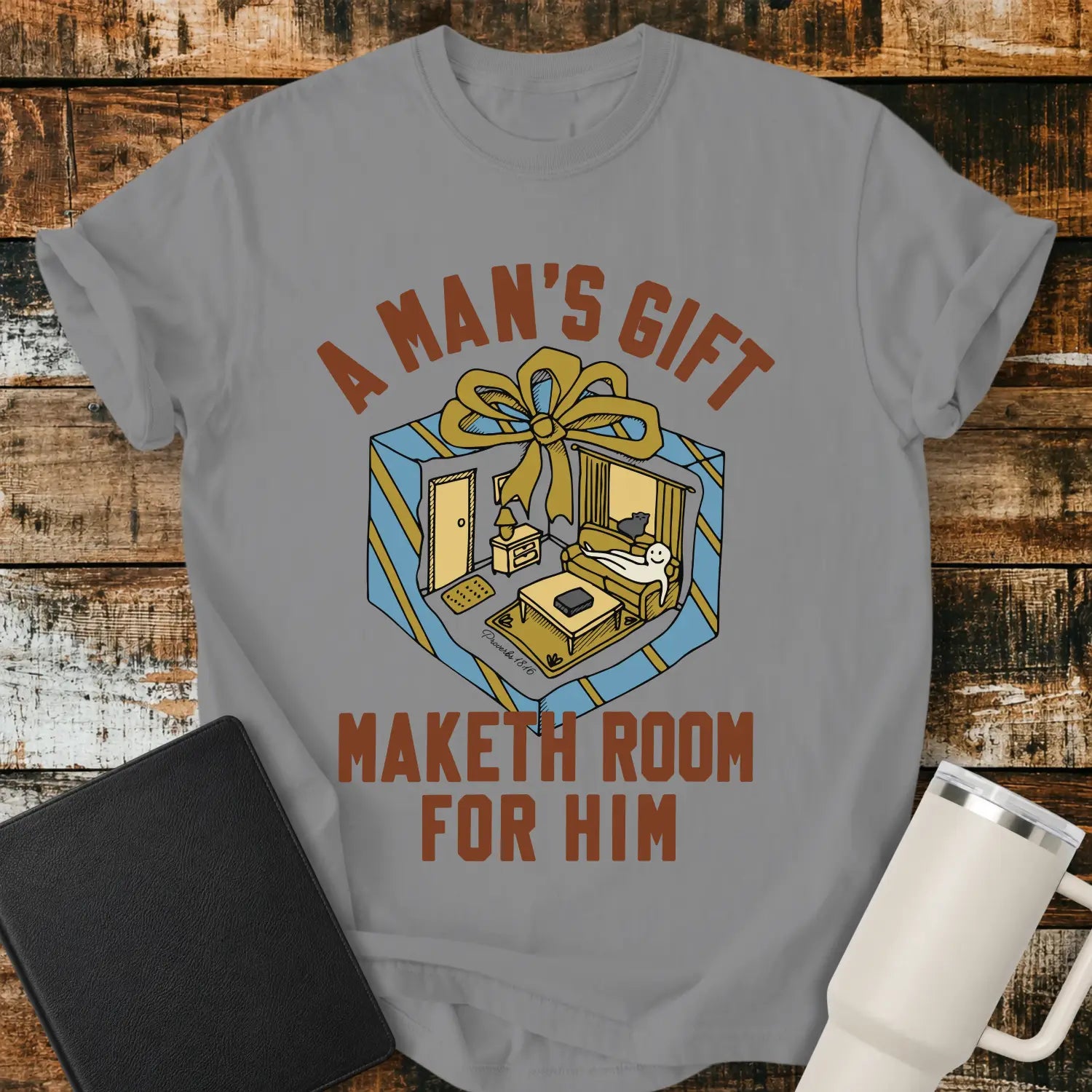 grey graphic T-shirt mockup featuring the phrase “A Man’s Gift Maketh Room for Him” in vintage-style lettering with an illustration of a gift box opening into a furnished room scene, styled with a black notebook and white tumbler on a rustic wood background.