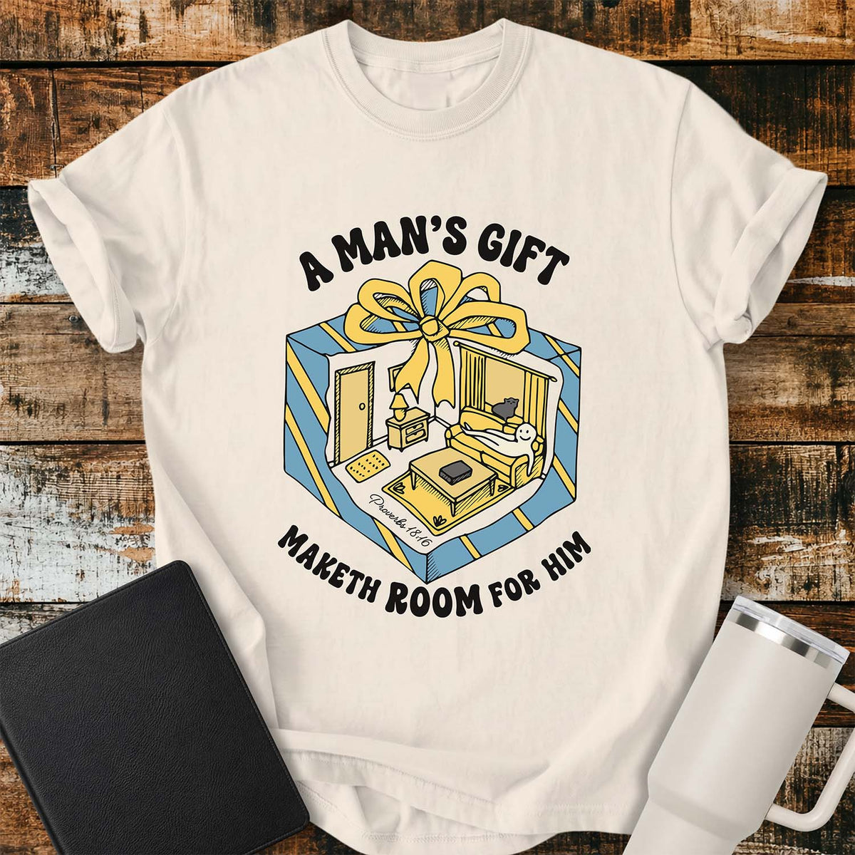ivory flat-lay t-shirt mockup featuring a colorful retro illustration of a gift box opened into a cozy living room with a sofa, lamp, coffee table, door, and window beneath a large yellow bow. Curved black lettering reads, "A MAN'S GIFT" above and "MAKETH ROOM FOR HIM" below, inspired by Proverbs 18:16. The shirt is displayed on a rustic wood plank background with a black leather Bible and a white insulated travel mug.
