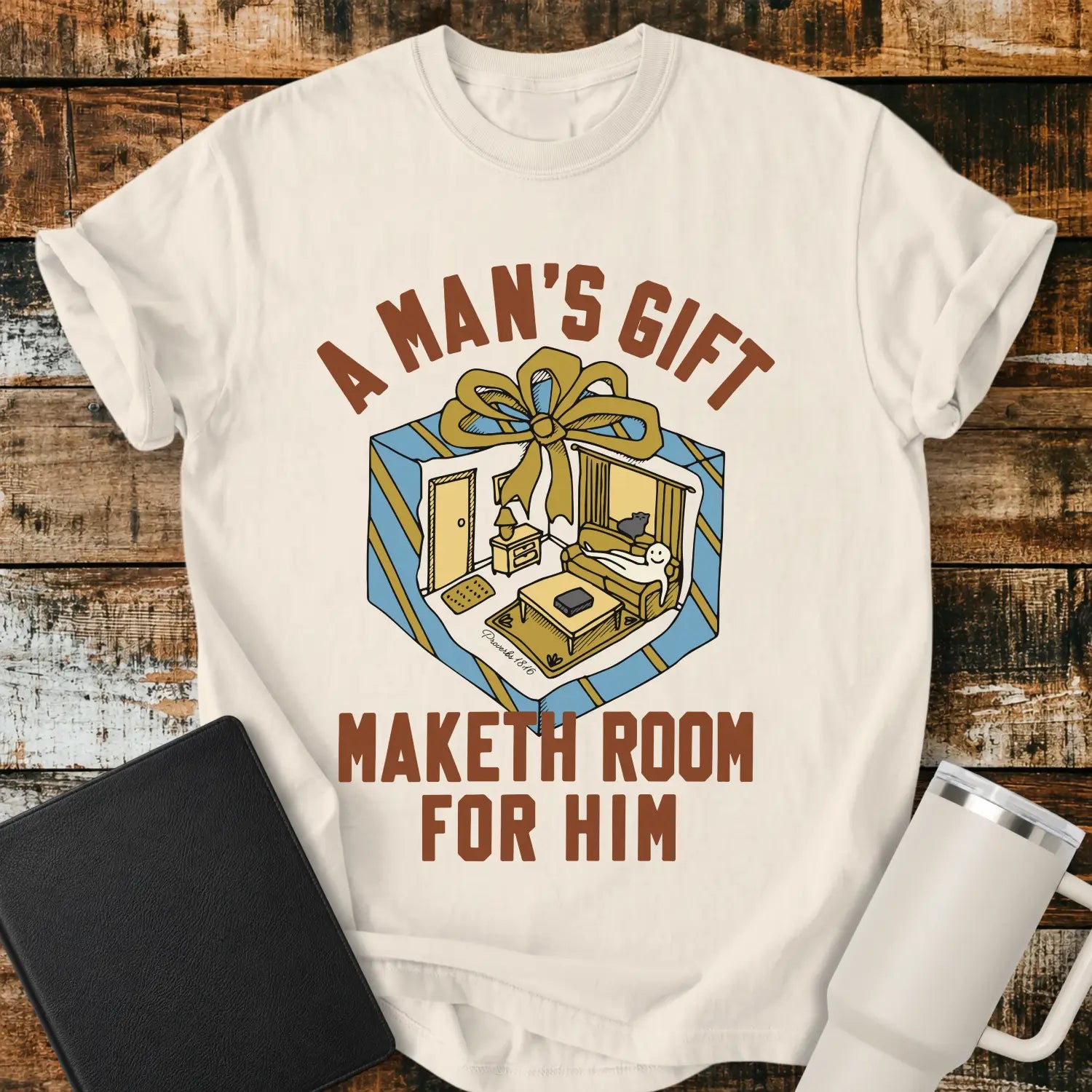 ivory graphic T-shirt mockup featuring the phrase “A Man’s Gift Maketh Room for Him” in vintage-style lettering with an illustration of a gift box opening into a furnished room scene, styled with a black notebook and white tumbler on a rustic wood background.