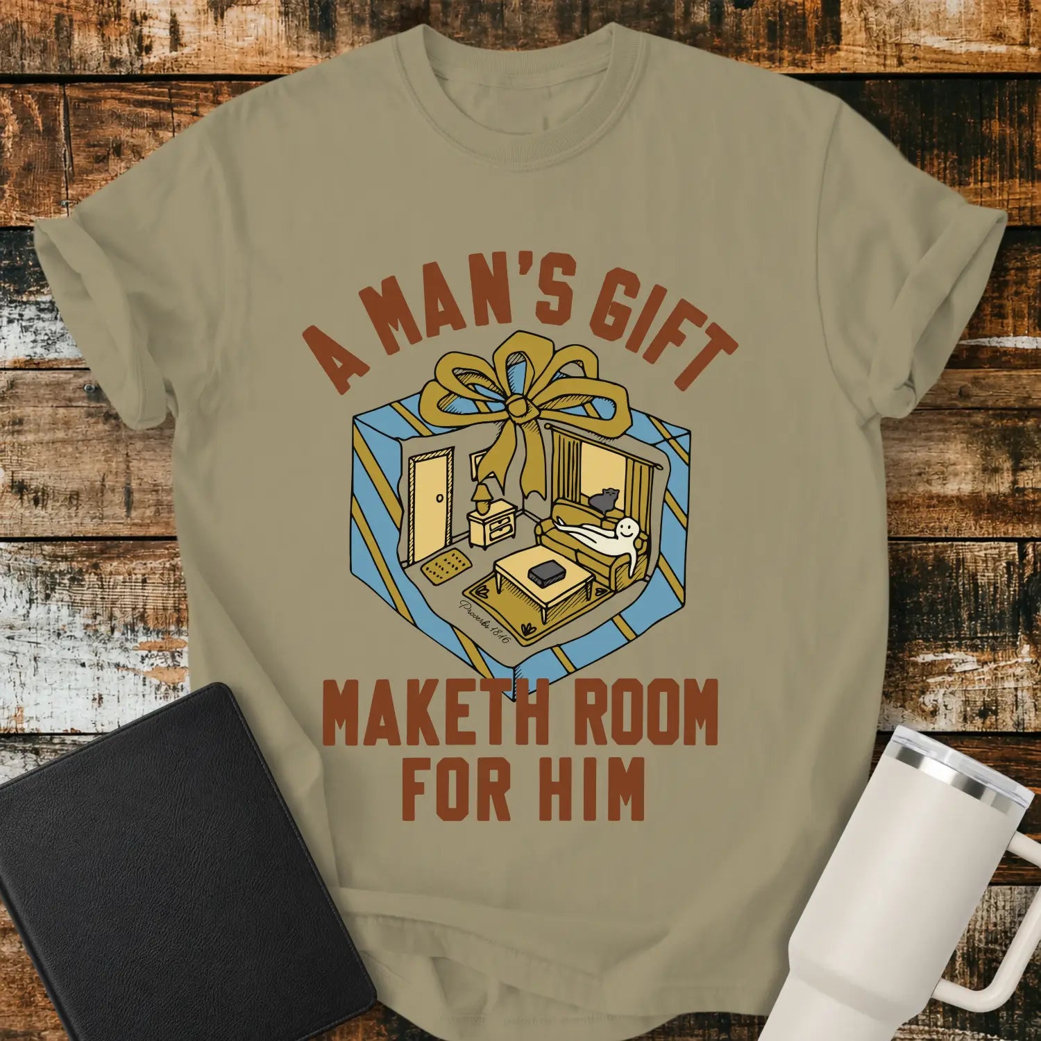 khaki graphic T-shirt mockup featuring the phrase “A Man’s Gift Maketh Room for Him” in vintage-style lettering with an illustration of a gift box opening into a furnished room scene, styled with a black notebook and white tumbler on a rustic wood background.